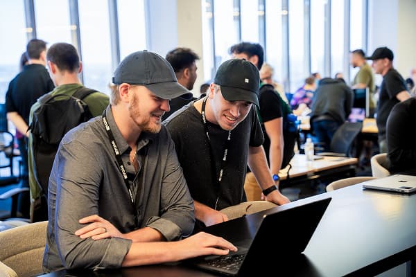 Two men in casual attire and baseball caps engage with a laptop, smiling. They're in a bright, busy office with others collaborating in the background.