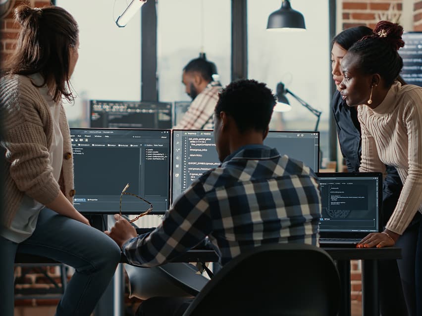 A diverse group of four people collaboratively work on laptops displaying code in a modern office. They appear focused and engaged in problem-solving.