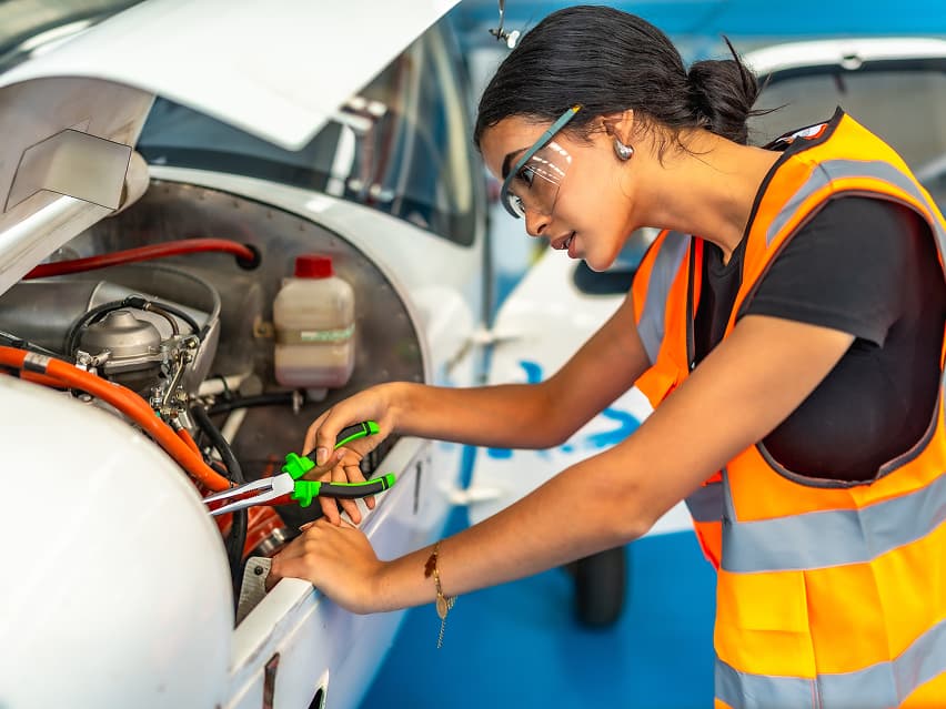 A woman in a safety vest and goggles is working on an aircraft engine using tools. She appears focused and intent in a hangar environment.