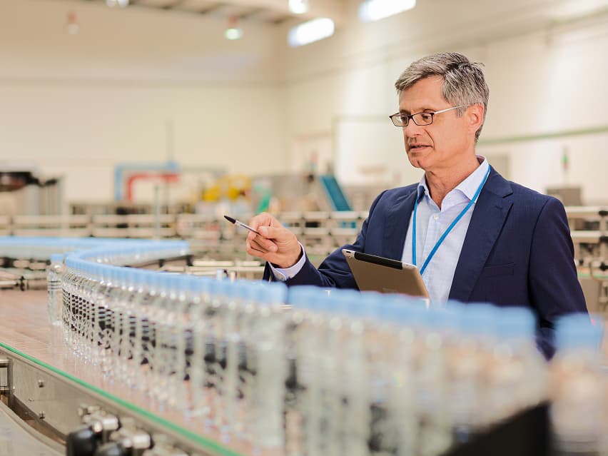 Man in a suit with glasses holds a tablet and pen, inspecting a row of bottled products on a factory conveyor belt, conveying focus and professionalism.
