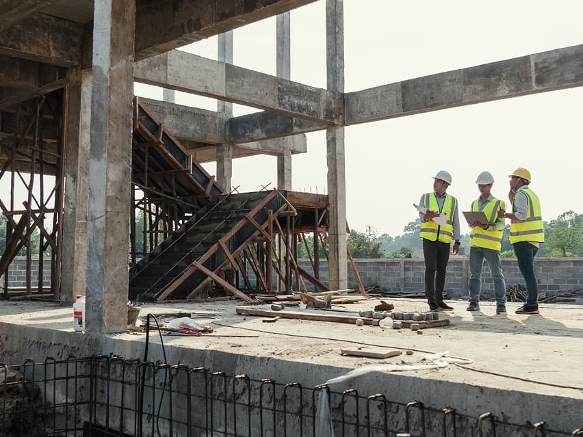Three construction workers in reflective vests and hard hats stand on a building site, discussing plans. Visible structures include concrete beams and a staircase.